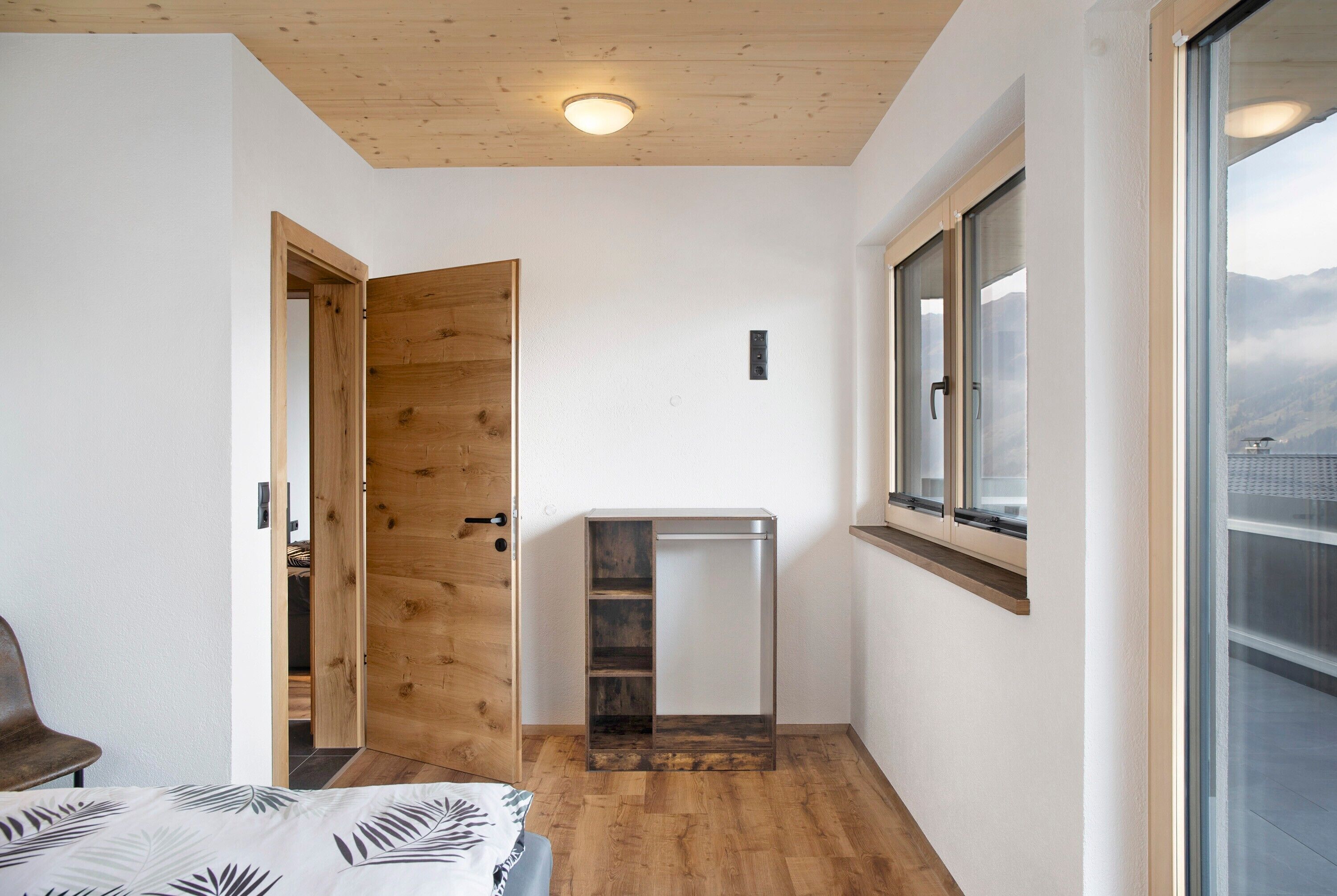 Un dormitorio con puerta de madera, cama con colcha de estampado de hojas, un escritorio pequeño y una ventana con vistas a las montañas.