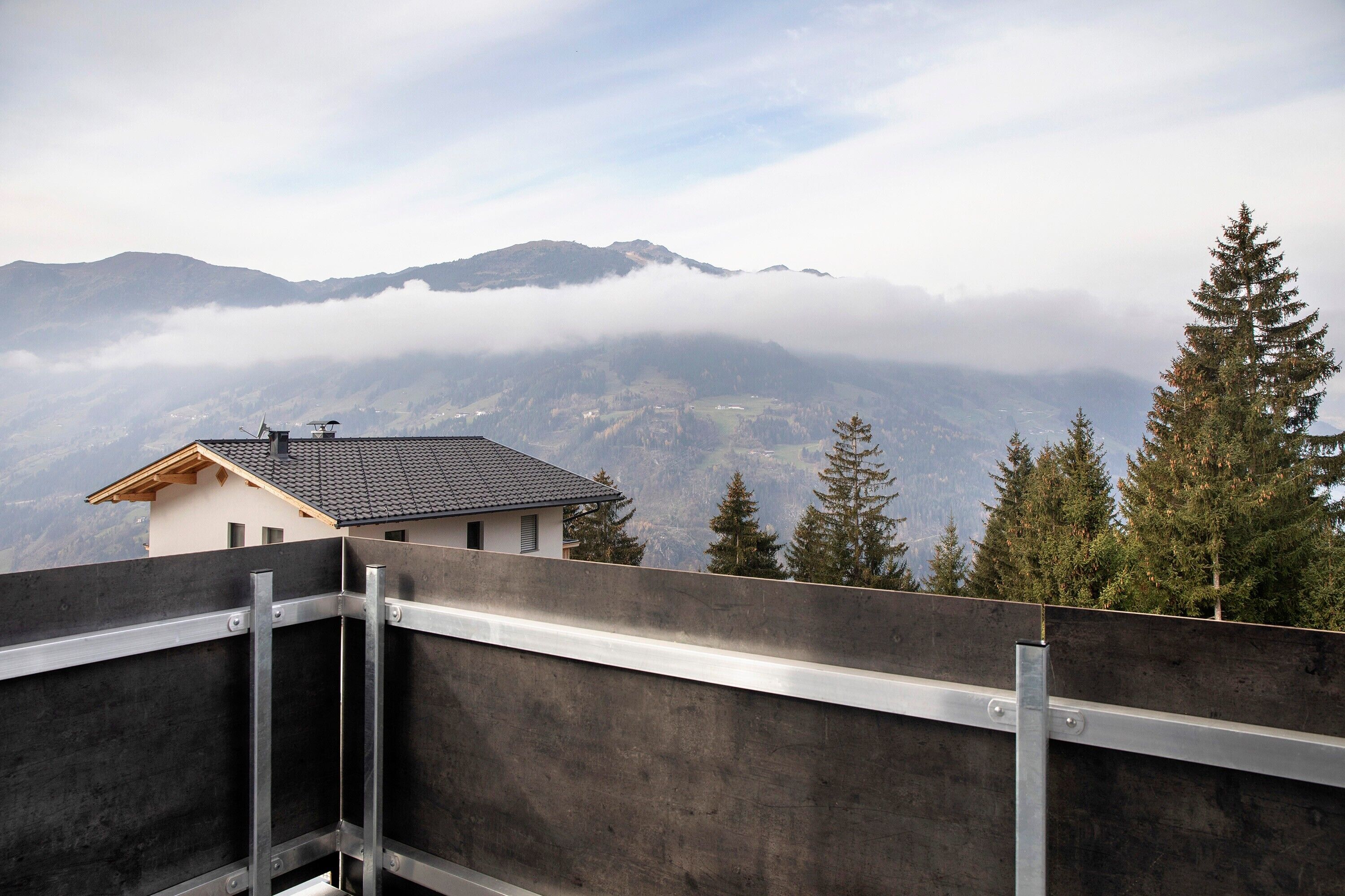 A building with a sloped roof, a metal railing, and a view of mountains and trees.