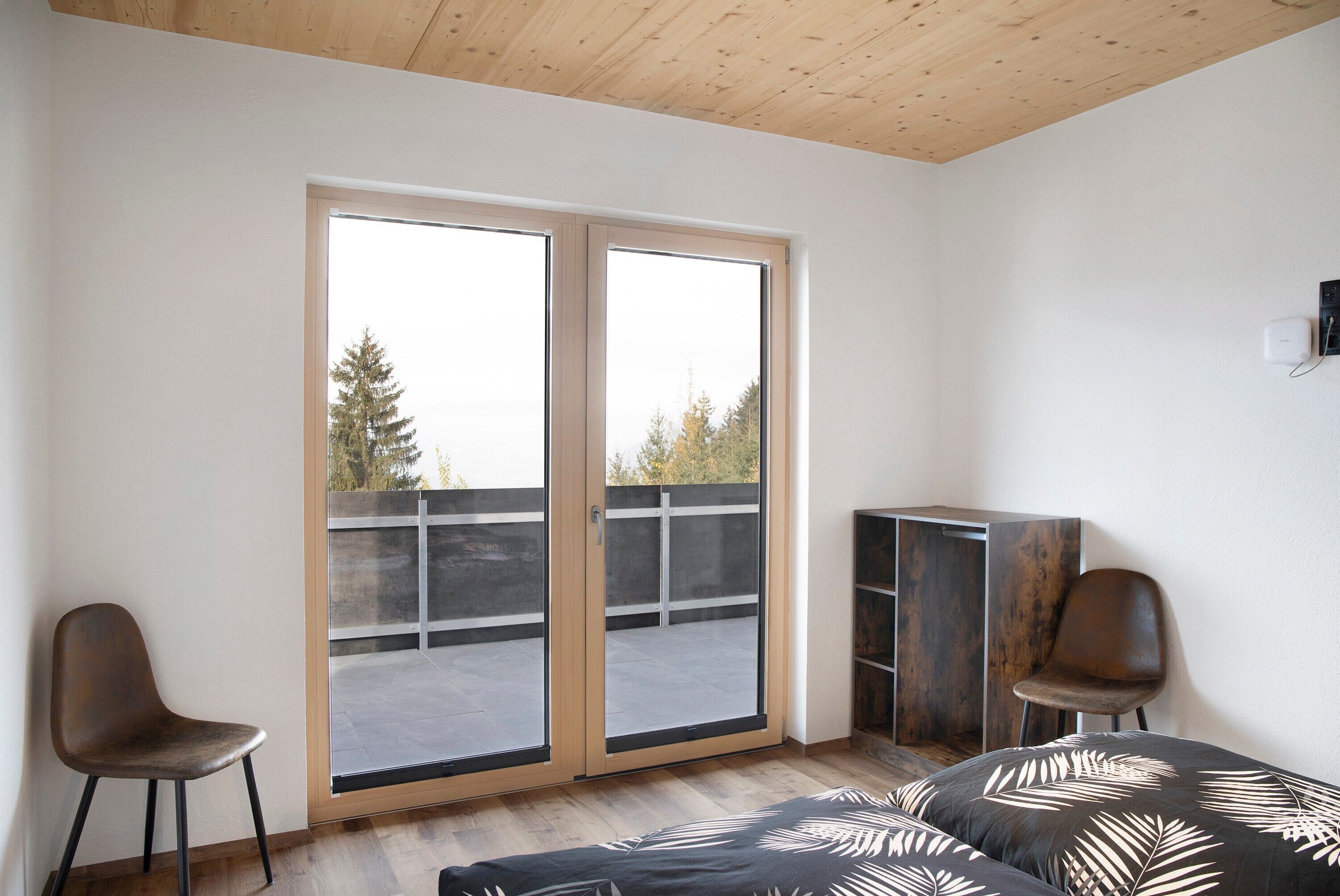 Een kamer met een grote schuifdeur naar een balkon, een bed met een beddegoed met een bladmotief, twee stoelen en een houten kast.