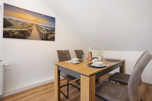 Uma área de refeições com uma mesa de madeira, quatro cadeiras, um vaso com planta e uma pintura de uma paisagem ao pôr do sol.