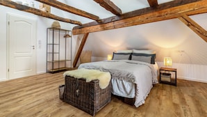 A bedroom with exposed wooden beams, a bed with patterned bedding, a wicker chest, and a bedside table with a lamp.