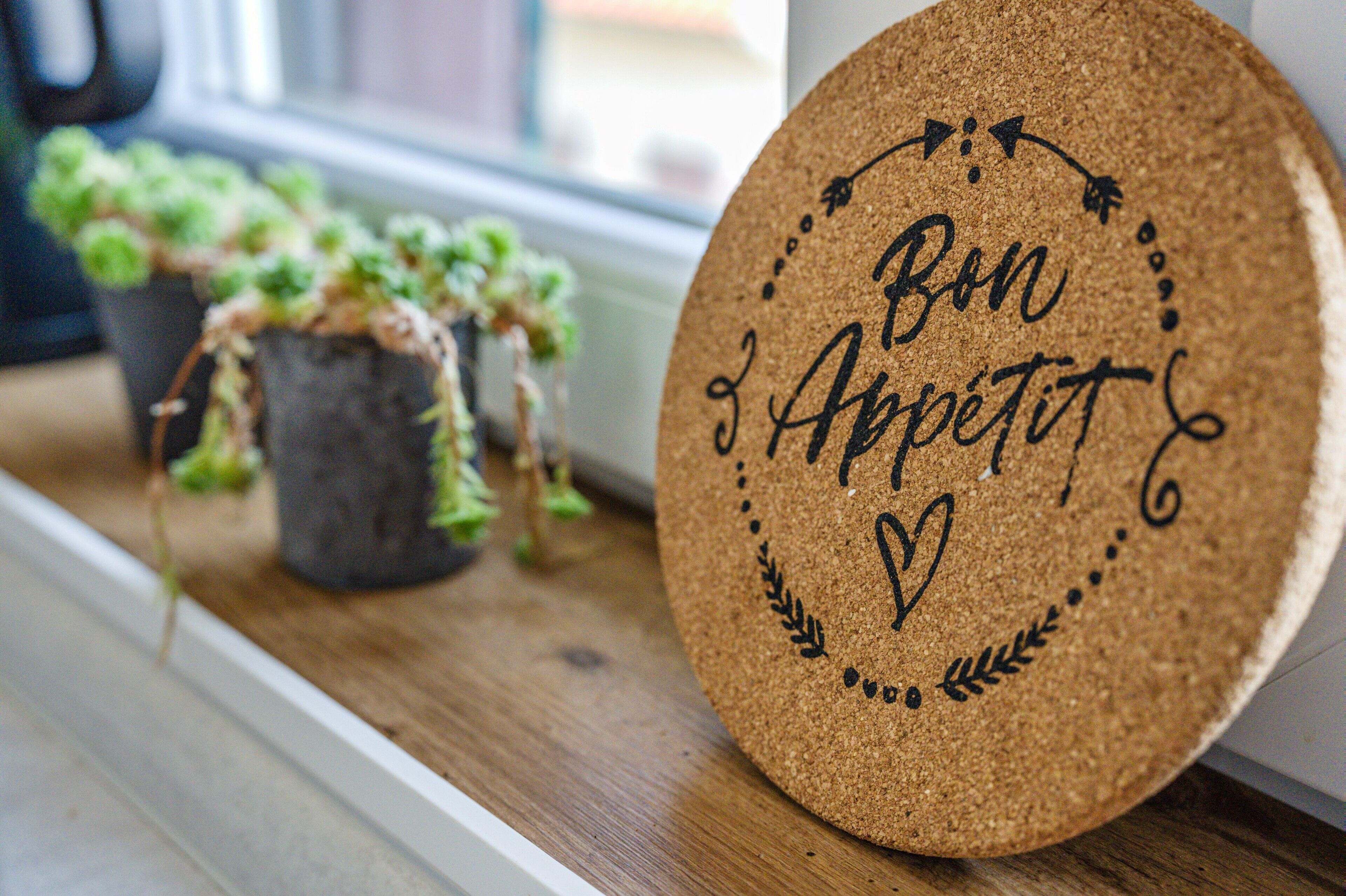 A corkboard with the words 'Bon Appetit' and decorative elements, placed on a wooden surface near potted plants.