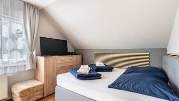 A bedroom with a bed, a television on a wooden cabinet, a small stool, and a window with sheer curtains.
