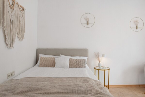 Una cama bien hecha con un cabecero gris, una mesita de noche con lámpara y adornos de macramé en la pared.