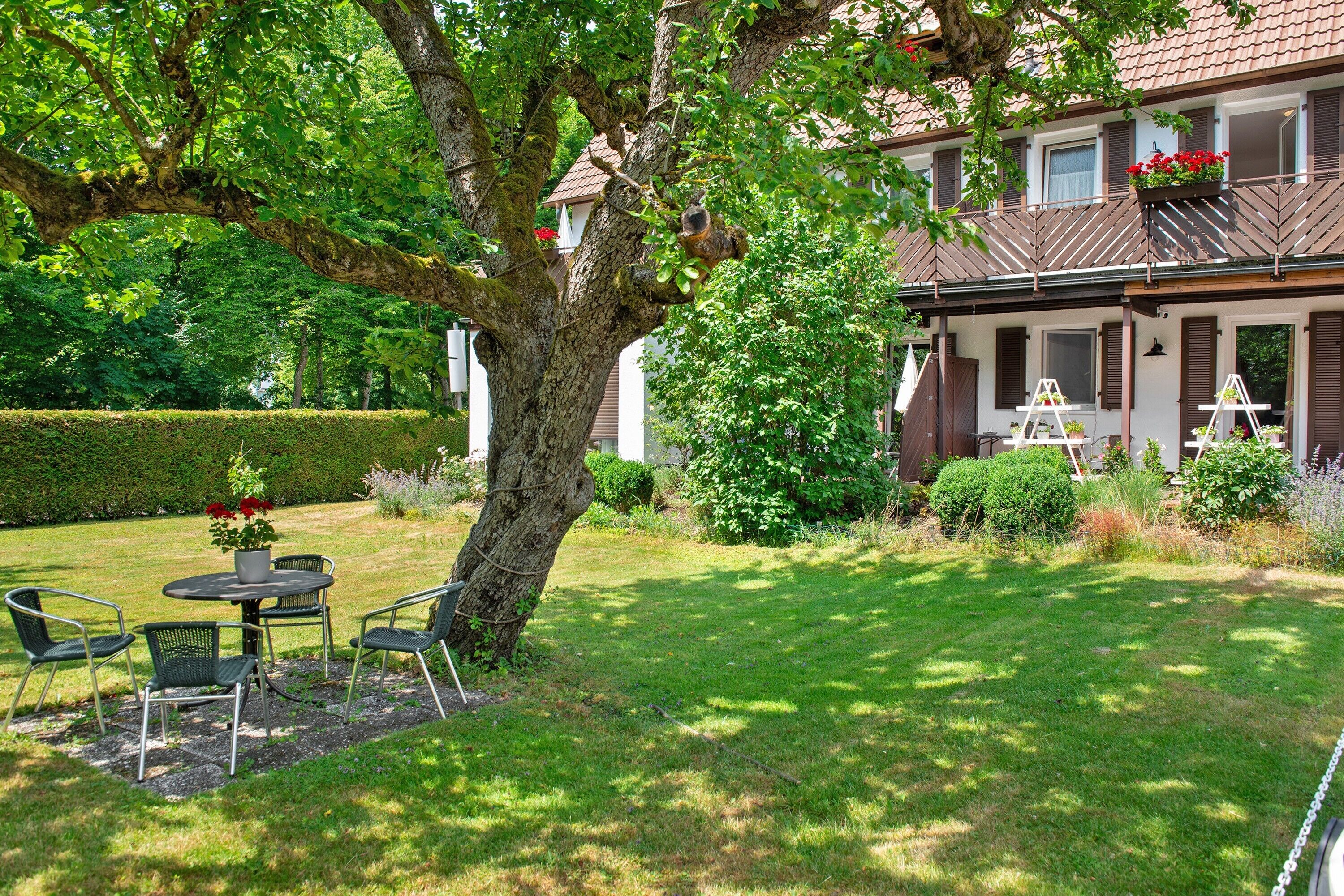 Een tuin met bomen, een patio met tafel en stoelen, een huis met balkon en een verzorgd gazon.