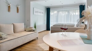 A modern living room with a sofa, a round table with glasses of red wine, and a view through a window with curtains. - Studio Apartment 'No. 1' with Shared Garden and Wi-Fi (Bad Dürrheim)