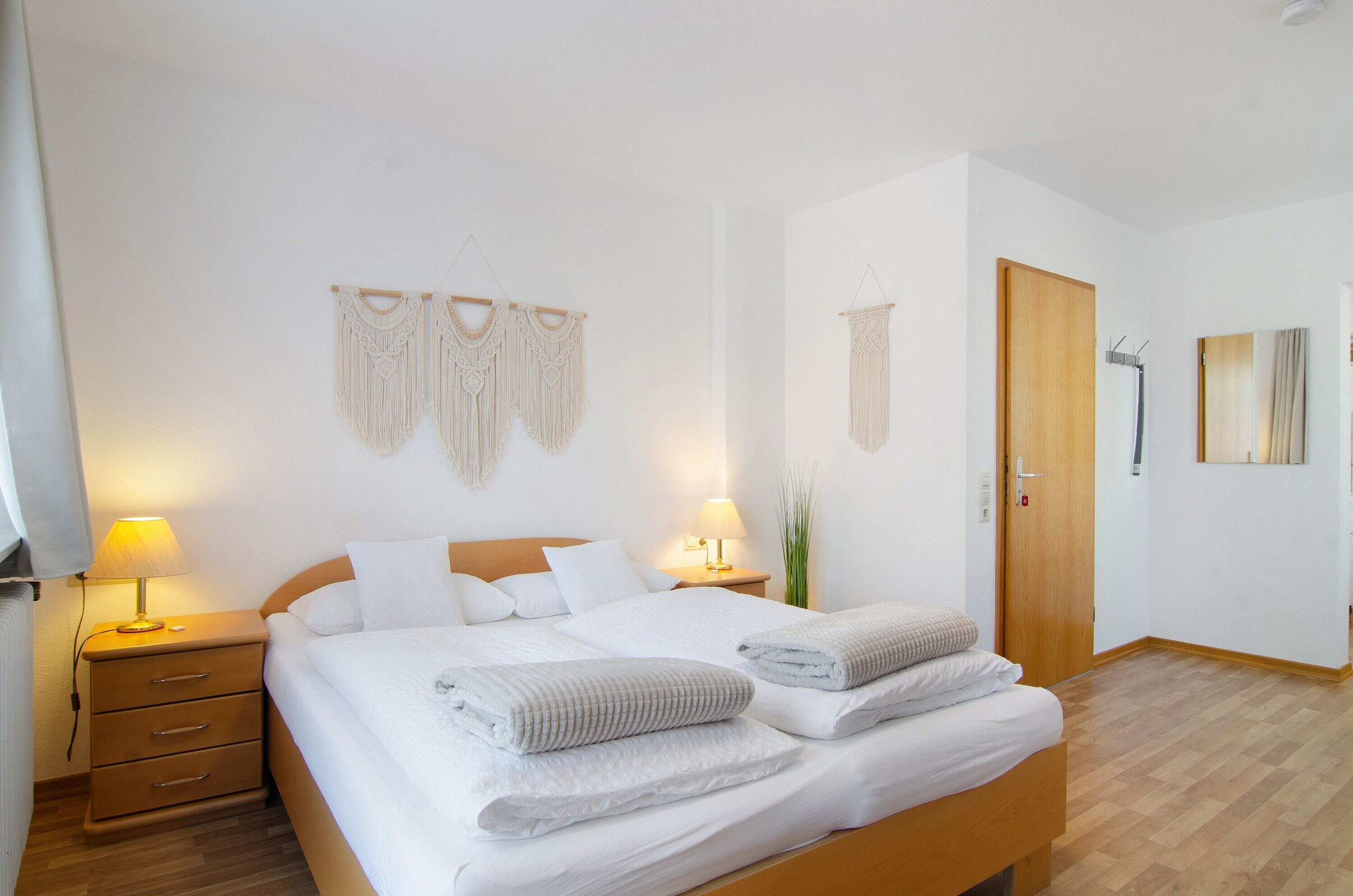 Un lit bien fait, avec du linge de lit et des serviettes blanches, une table de chevet en bois avec une lampe, une porte en bois et une décoration en macramé accrochée au mur.