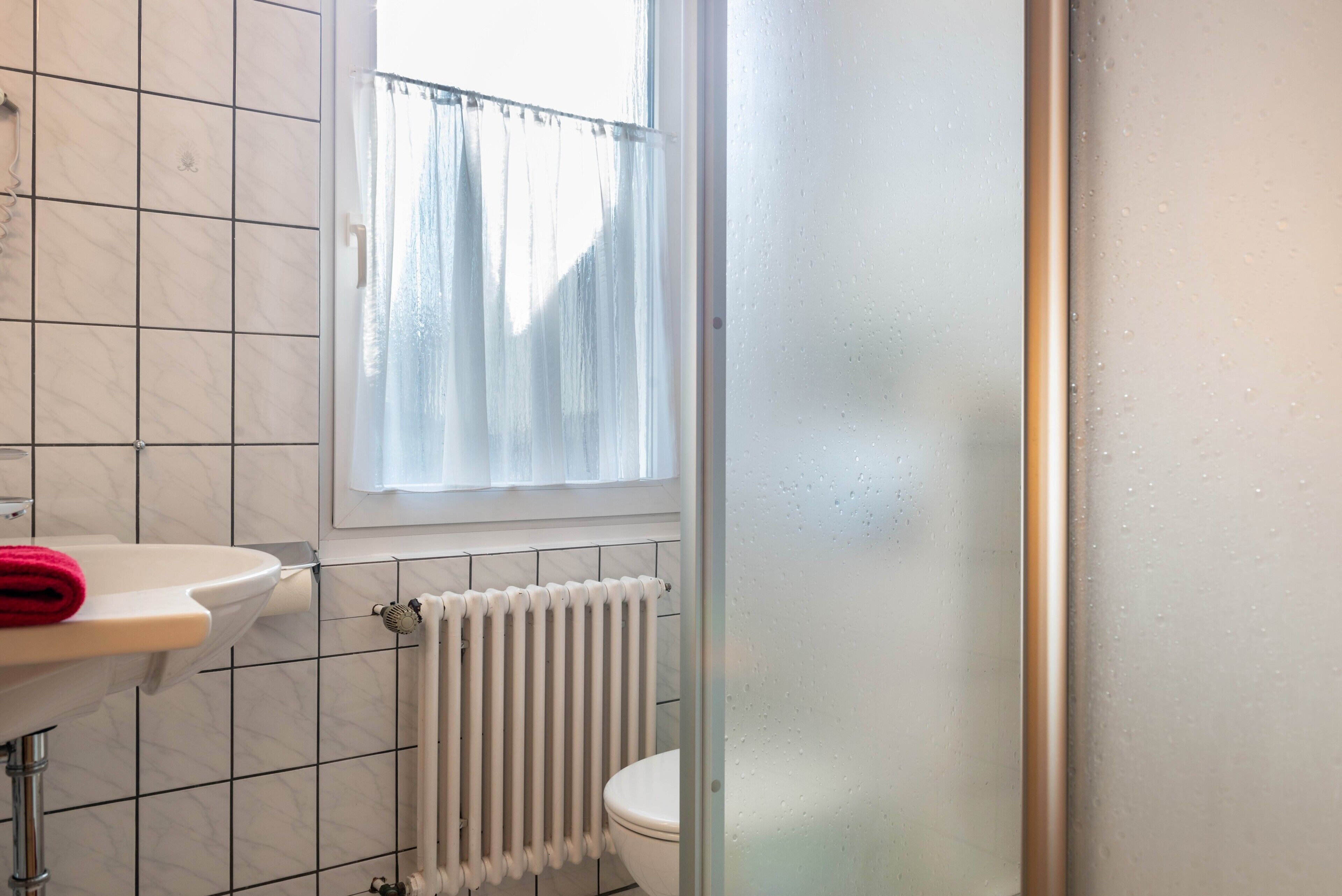 Une salle de bain avec un lavabo blanc, des toilettes, une fenêtre avec un rideau et un radiateur.