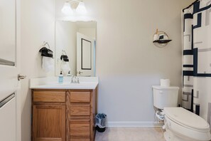 Un baño con tocador de madera, lavamanos blanco, espejo, inodoro y cortina de ducha con estampado geométrico.