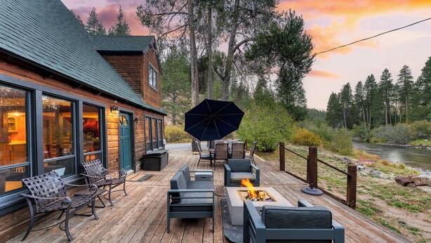 A wooden deck with outdoor seating, a fire pit, and a view of a river and forest. - Riversong Lodge by AvantStay | Sauna + Hot Tub | On The Truckee River (Truckee)