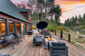 A wooden deck with outdoor seating, a fire pit, and a view of a river and forest. - Riversong Lodge by AvantStay | Sauna + Hot Tub | On The Truckee River (Truckee)