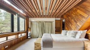 A bedroom with a large bed, wooden ceiling beams, a window with curtains, and a view of trees. - Riversong Lodge by AvantStay | Sauna + Hot Tub | On The Truckee River (Truckee)