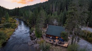 A cabin with a deck overlooking a river, surrounded by dense forest. - Riversong Lodge by AvantStay | Sauna + Hot Tub | On The Truckee River (Truckee)