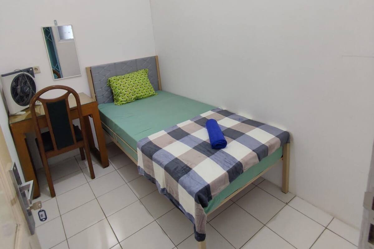 A small, clean room with a single bed, a chair, a desk with a mirror, and a fan.