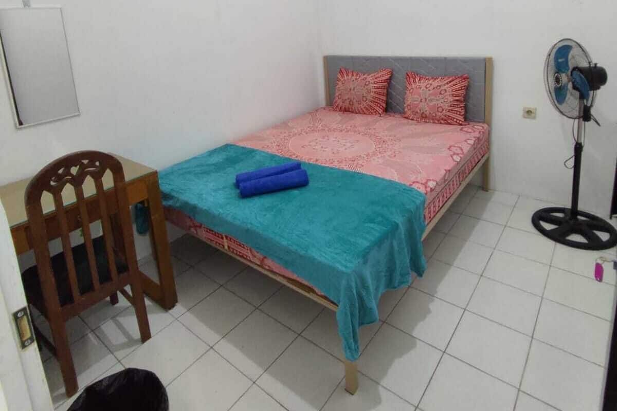A small, clean room with a single bed, a chair, a desk, and a fan.