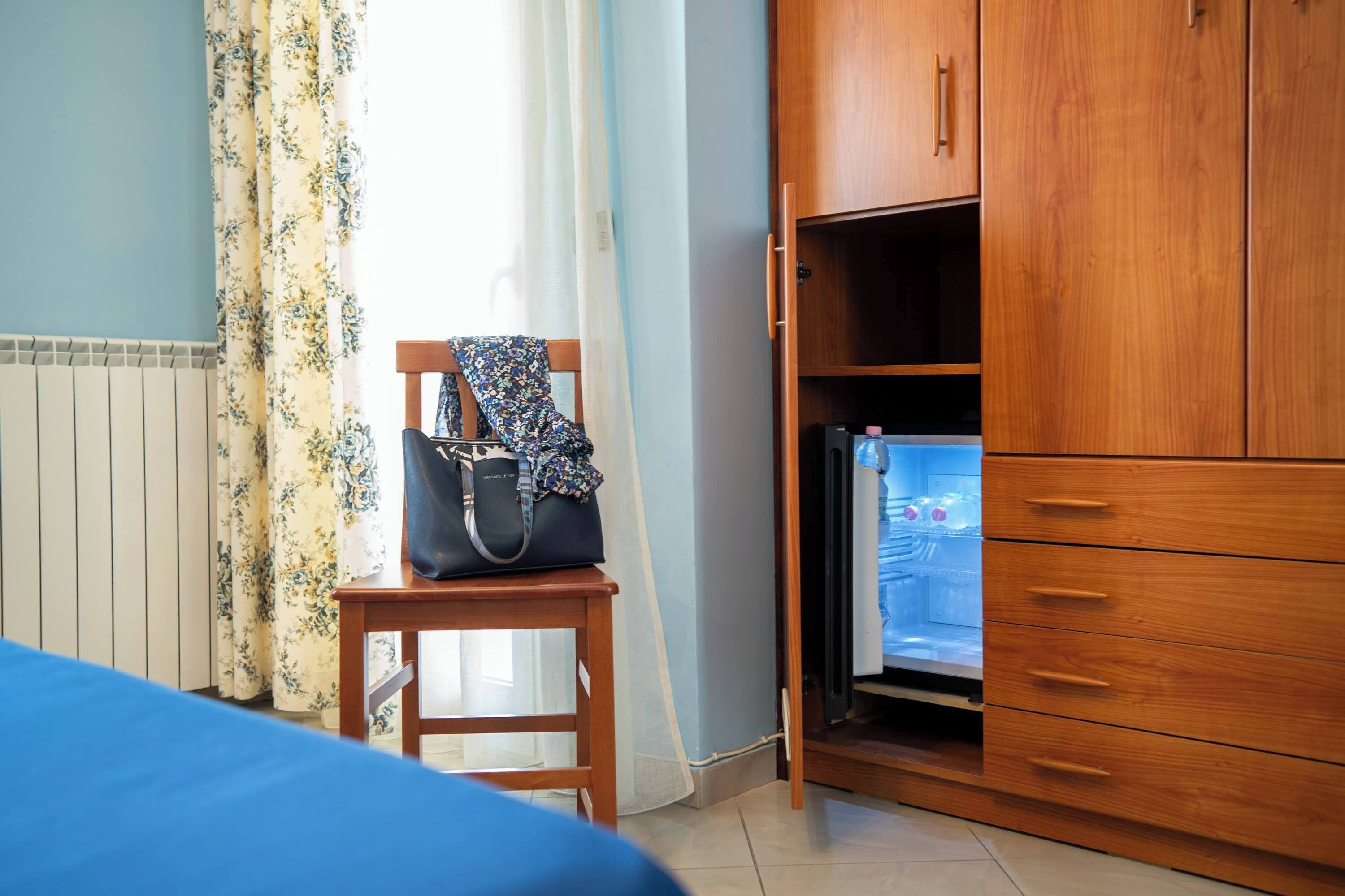 Una habitación con cama, un armario de madera, una mesita con un bolso y un refrigerador.