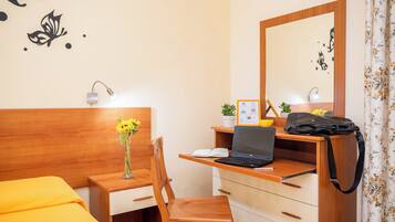 Habitación de hotel con un escritorio de madera, una computadora portátil, un jarrón con flores, una cama con colcha naranja, un espejo y una cortina con motivos florales.