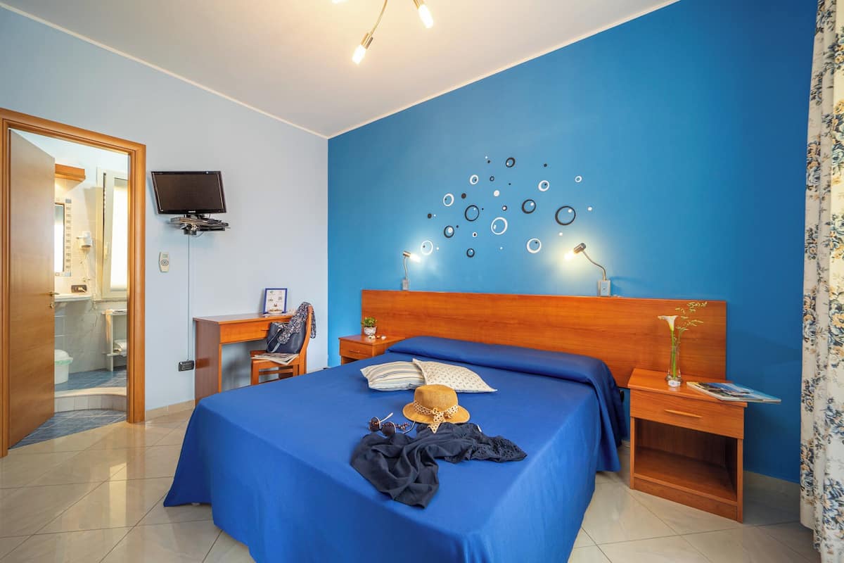 A hotel room with a blue wall, a wooden bed, a desk, a television, and a bathroom visible through an open door.