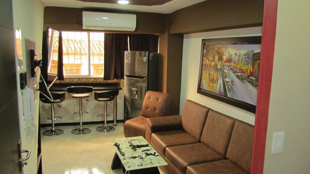 A compact living space with a brown sofa, a small table with a decorative item, a bar area with stools, a refrigerator, and a large painting on the wall. - One Bedroom AC Hot Tub 1 block Parque Poblado (Medellin)