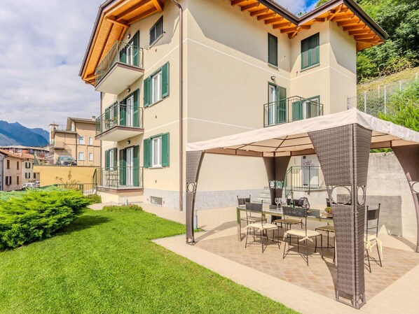 Un immeuble à plusieurs étages doté d’un espace extérieur couvert avec des sièges, d’une pelouse bien entretenue et d’un paysage montagneux en arrière-plan.