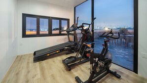 Un gimnasio con caminadora, elíptica y bicicleta estática, ventanas amplias con vista a la ciudad y piso de madera.