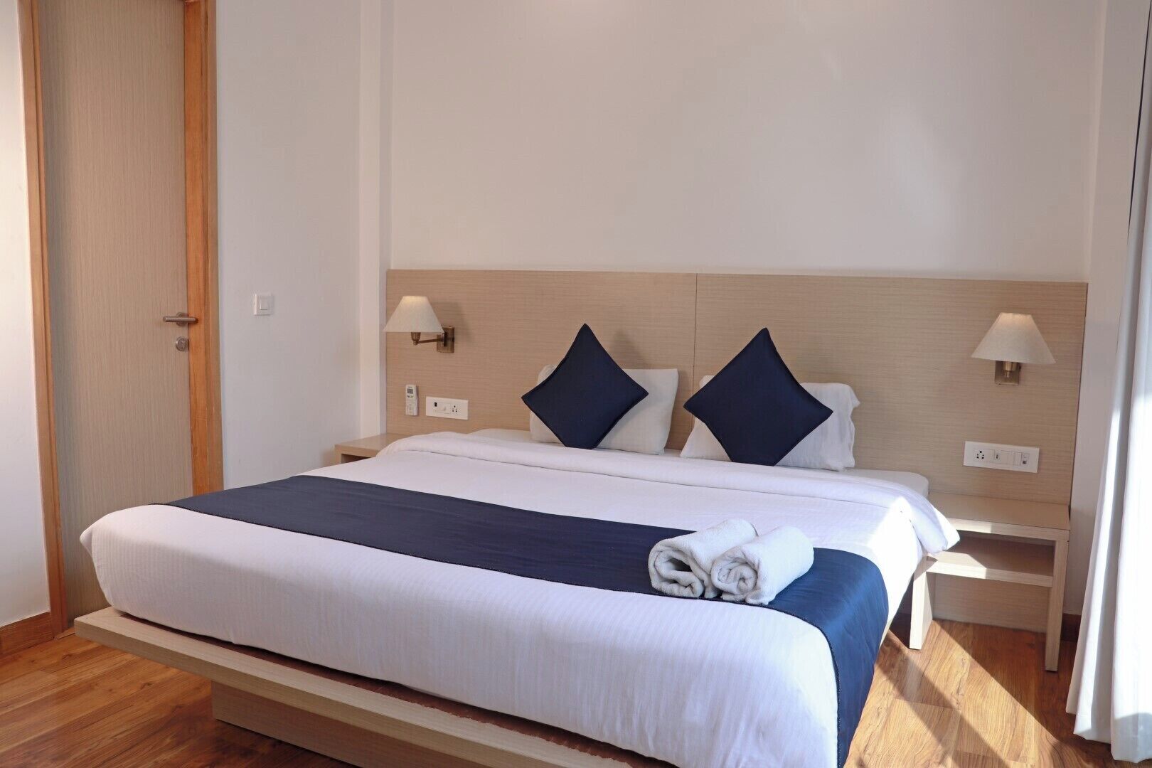 Una cama bien tendida con colcha azul y blanca, dos almohadas y una toalla enrollada al lado. Hay dos mesitas de noche con lámparas, un cabecero de madera y un suelo de madera.