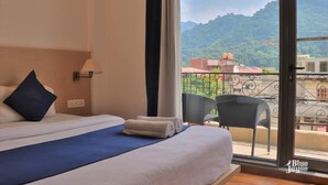 A hotel room with a bed, a chair, and a view of a mountain and buildings. - The Blue Jay Hostel | Dorms & Rooms (Rishikesh)