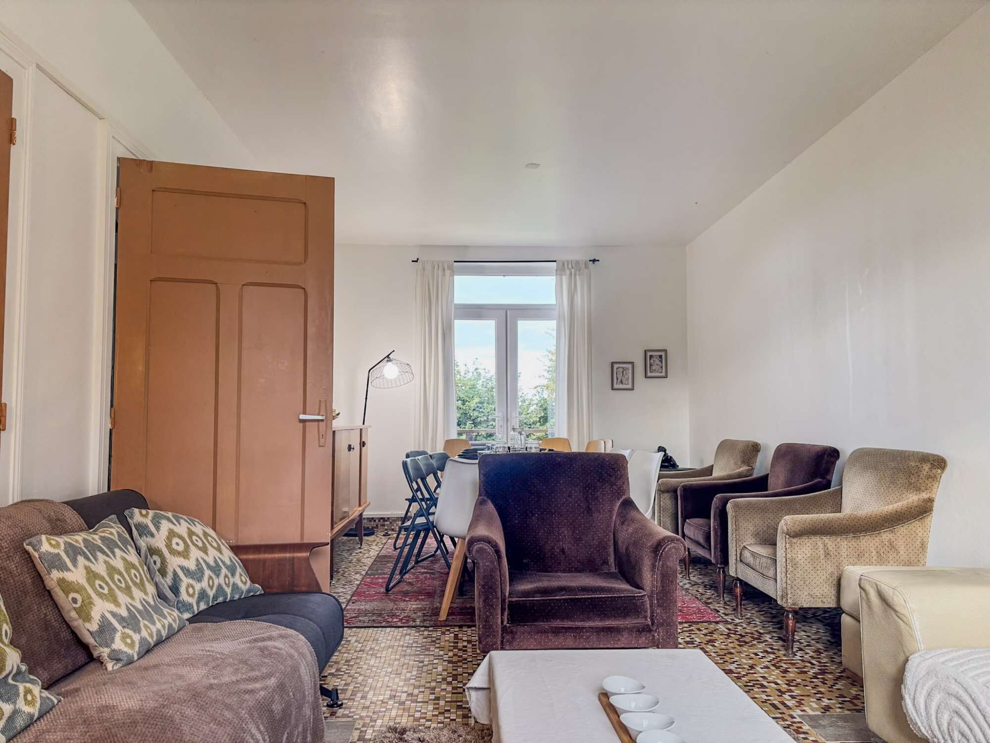 A living room with a sofa, armchairs, a dining table with chairs, a large window, and a door.