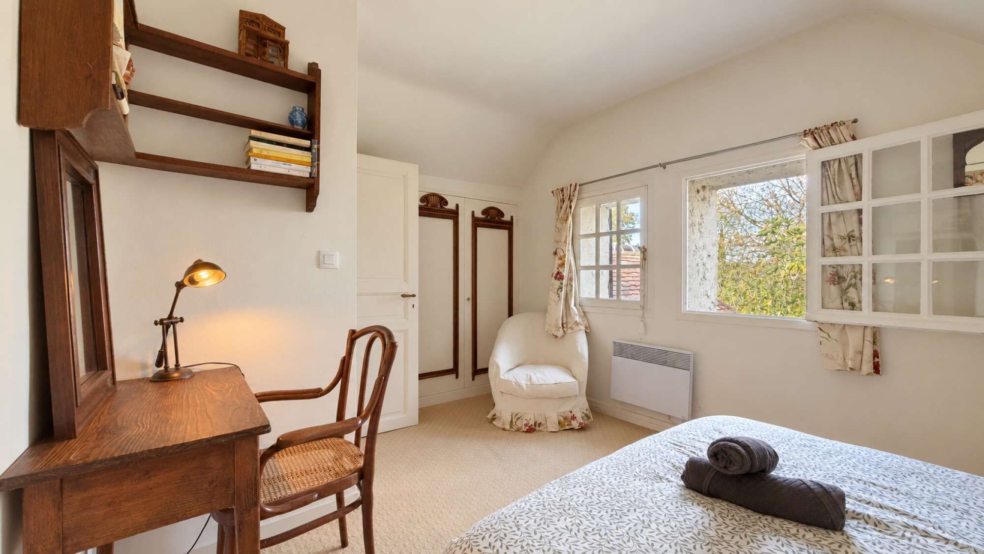 A bedroom with a bed, a chair, a desk with a lamp, a window with curtains, and a radiator.