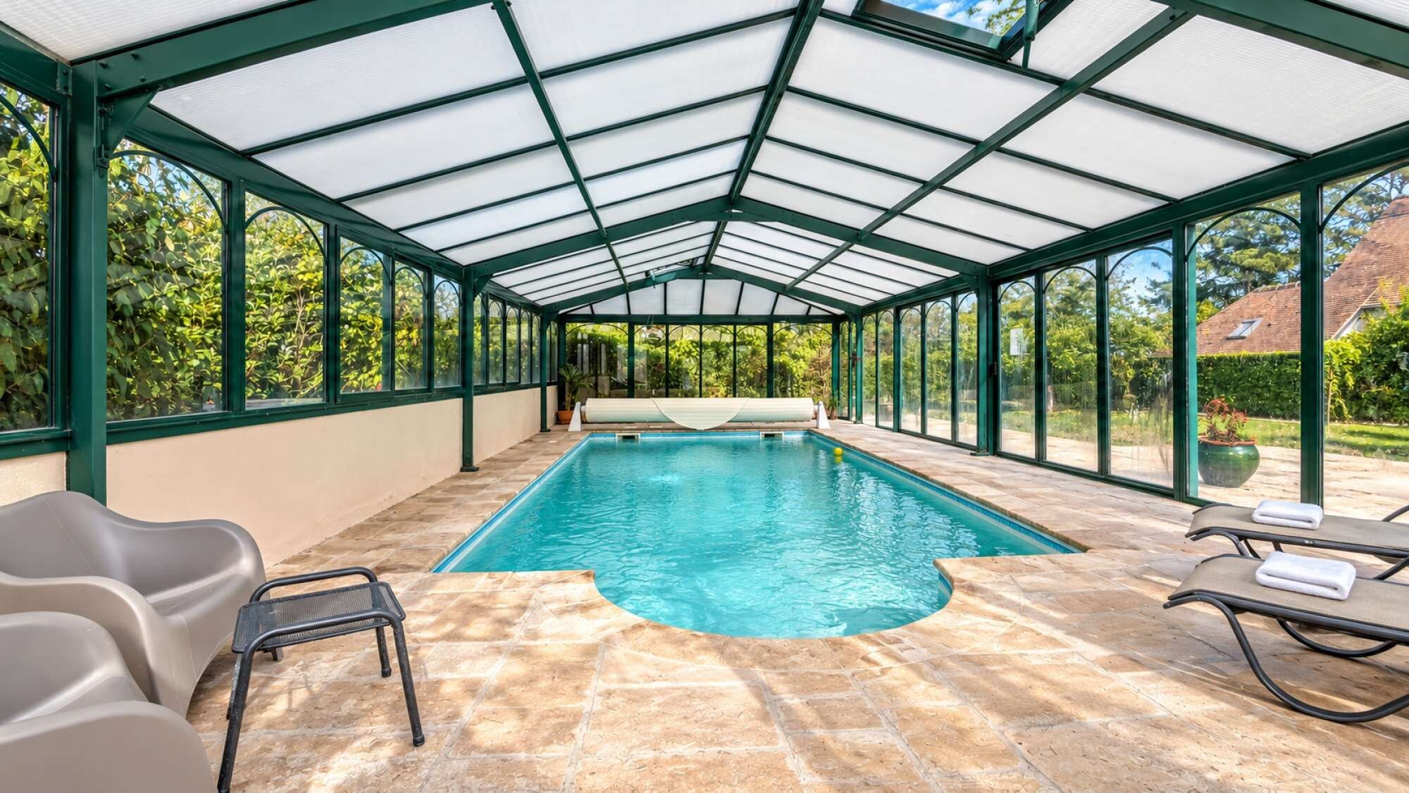 Une piscine intérieure avec un toit en verre, un sol en pierre et des chaises longues.