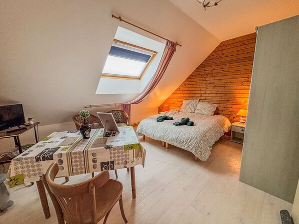 Une pièce chaleureuse avec un plafond en pente, un lit recouvert d’une couverture à motifs, une table avec un ordinateur portable et une petite plante.