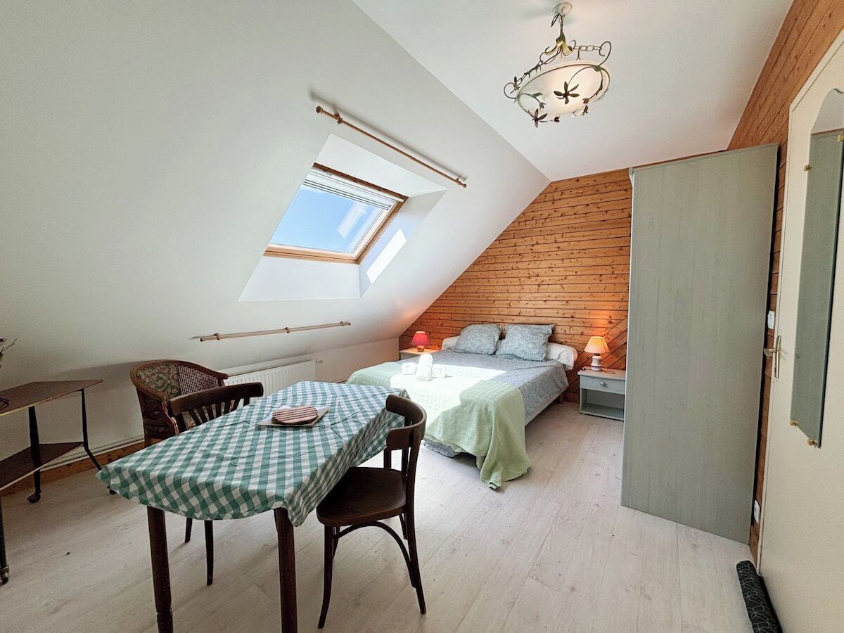 Ett mysigt sovrum på vinden med ett sluttande tak, ett takfönster, en säng med ett grönt överkast, ett litet bord med en rutig duk och en stol.
