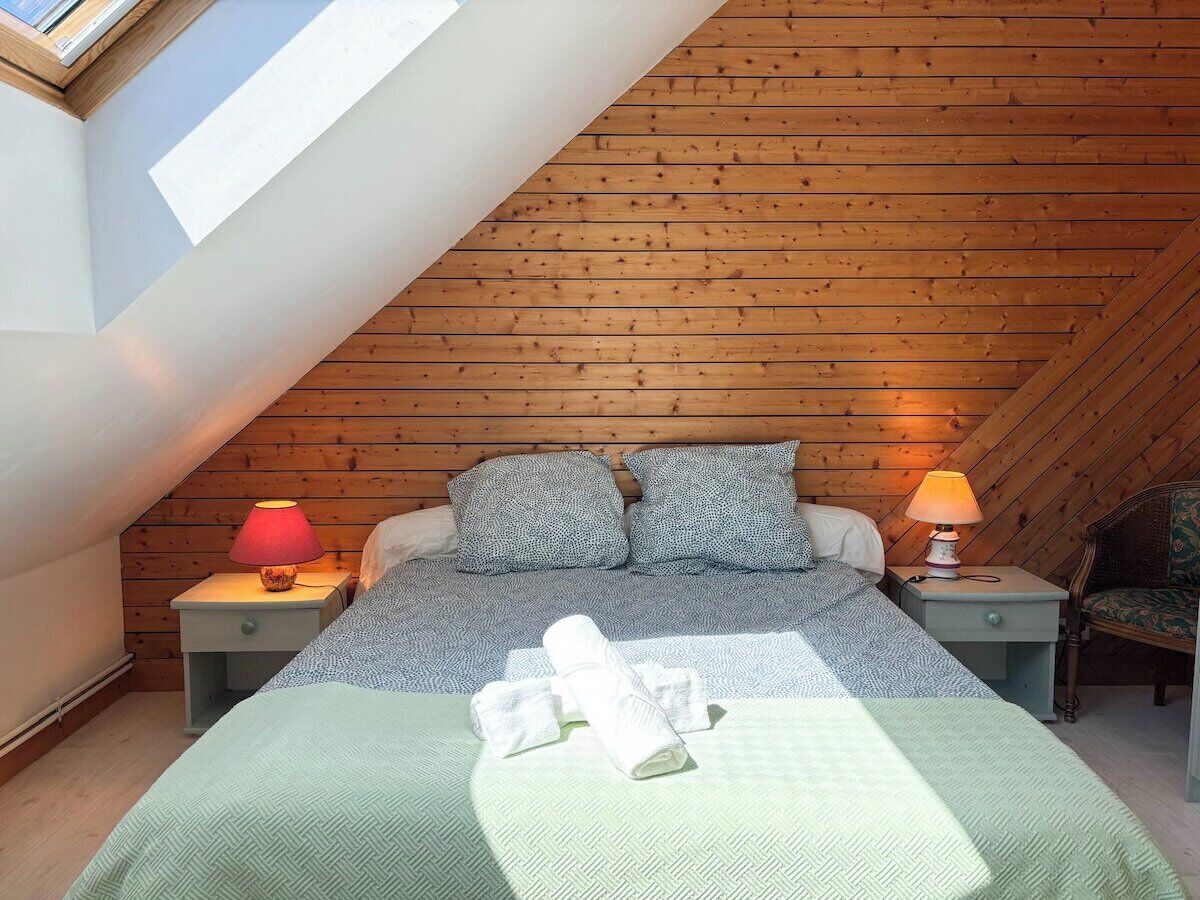A cozy attic bedroom with a sloped ceiling, wooden walls, a bed with patterned bedding, two bedside tables with lamps, and a window providing natural light.