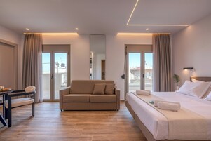 A modern hotel room with a sofa, a bed, a dining table, and a view of the city through large windows.