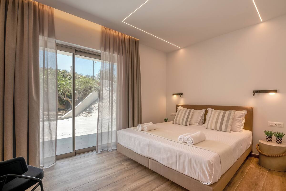 A modern bedroom with a large bed, a sliding glass door to a balcony, and a small round table with a plant.