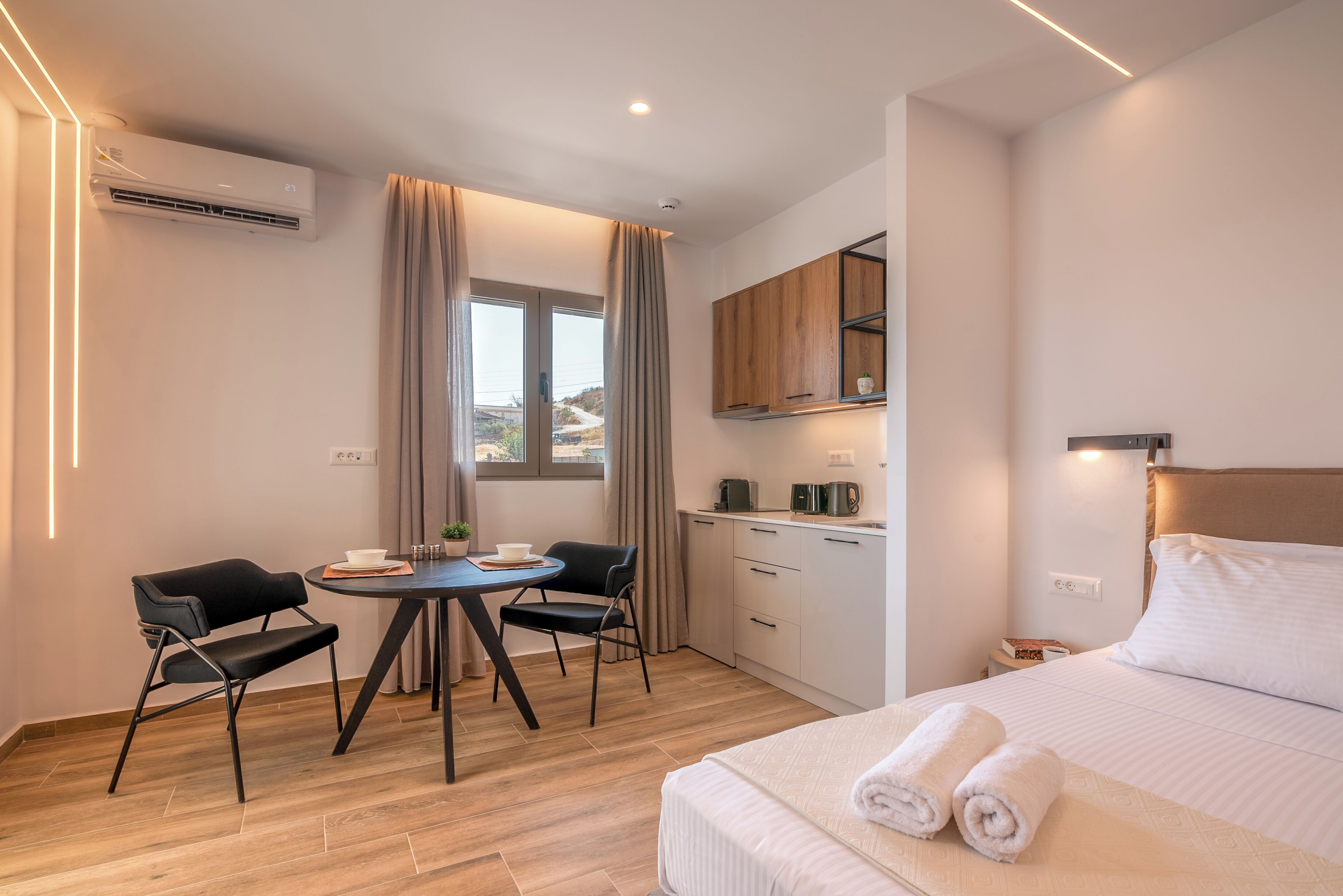Une chambre d’hôtel moderne comprenant un lit, une table à manger avec des chaises, un coin cuisine et une grande fenêtre avec des rideaux.