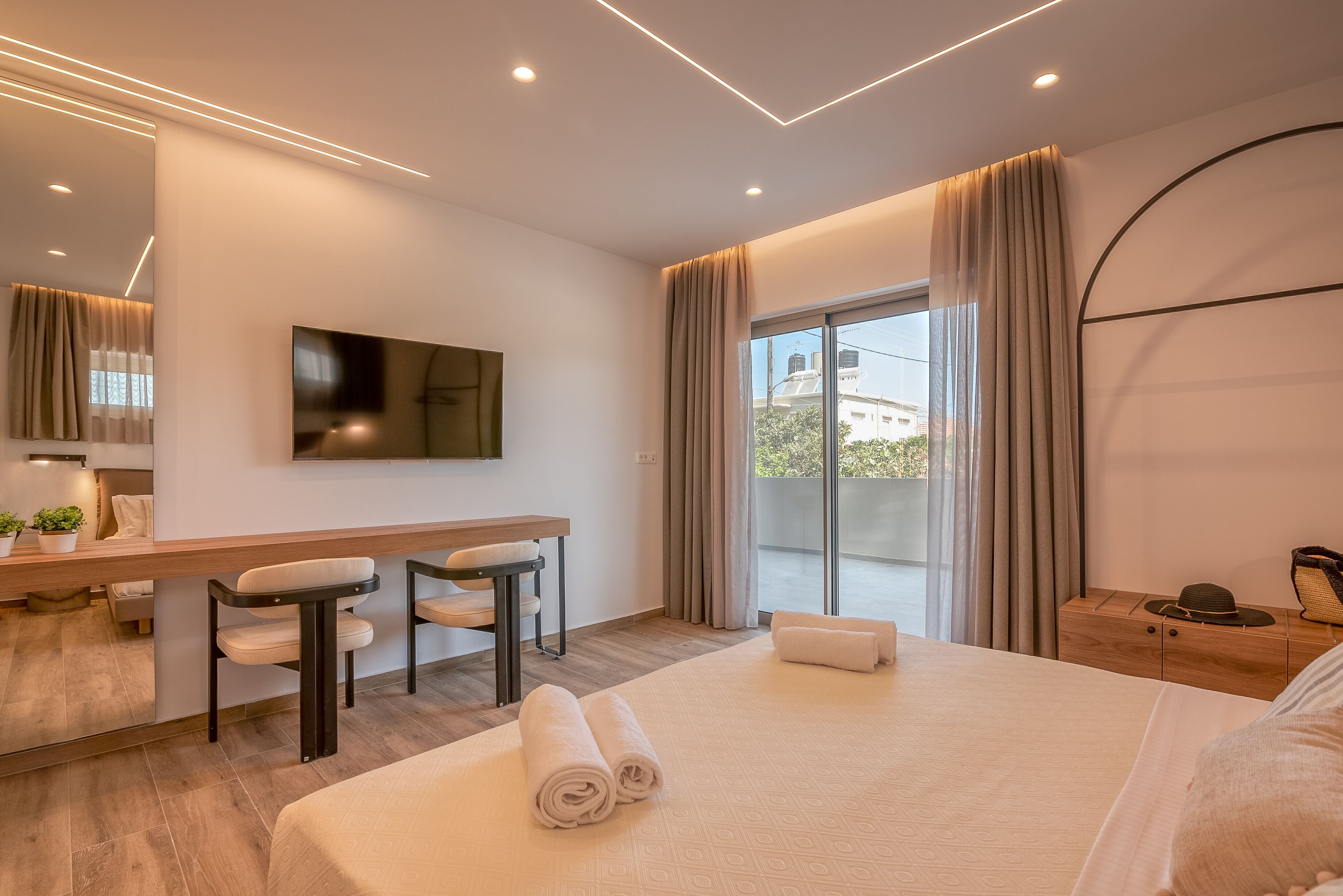 A modern hotel room with a large bed, a desk with chairs, a flat-screen TV, and a balcony with curtains.