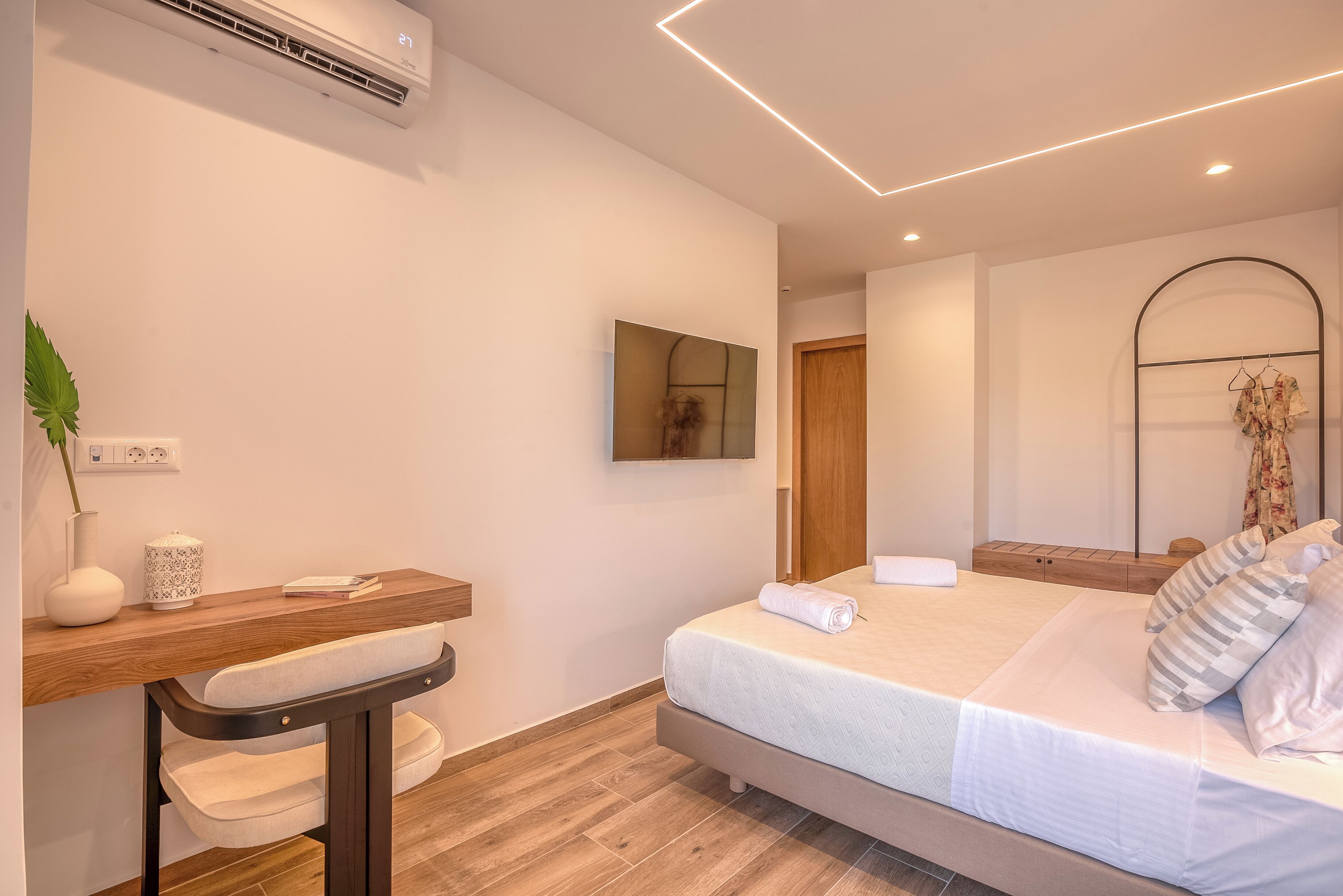 A modern bedroom with a wooden desk, a bedside table, a bed with white bedding, and a mirror on the wall.