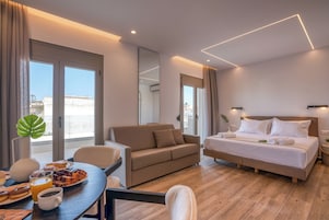 Una habitación de hotel moderna con cama, sofá, una mesa de comedor con desayuno y ventanas grandes.