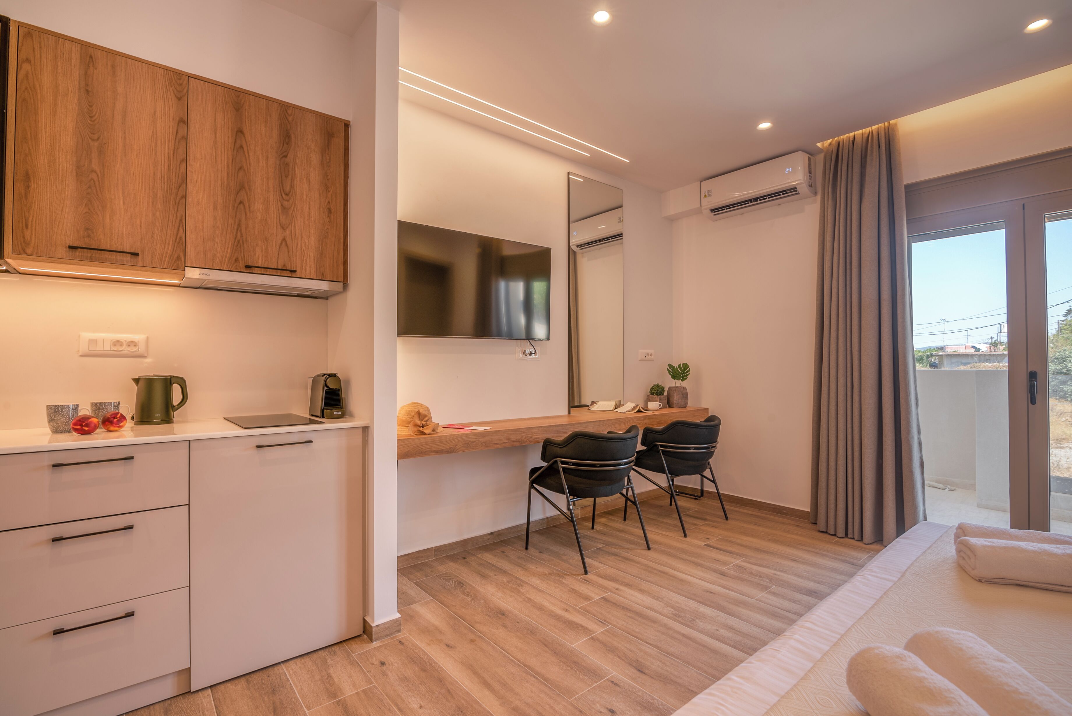 Una cucina moderna con mobili in legno, un piano di lavoro con un bollitore e uno specchio sopra il lavello. Ci sono due sedie nere e un tavolino con una pianta.