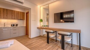 A modern kitchen with wooden cabinets, a built-in oven, and a coffee machine. There is a dining area with a wooden table and two chairs. A large mirror and a flat-screen TV are mounted on the wall. - kiwi butique studios (Kissamos)