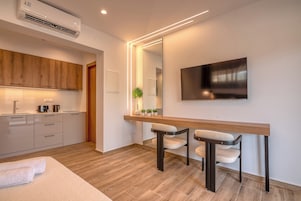 A modern kitchen with wooden cabinets, a built-in oven, and a coffee machine. There is a dining area with a wooden table and two chairs. A large mirror and a flat-screen TV are mounted on the wall.