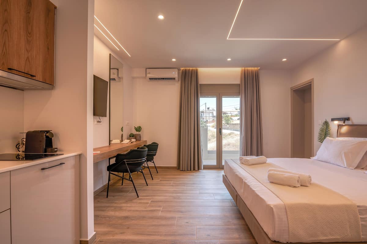 A modern hotel room with a wooden desk, a bed with white linens, a small seating area, and a view through the glass door.