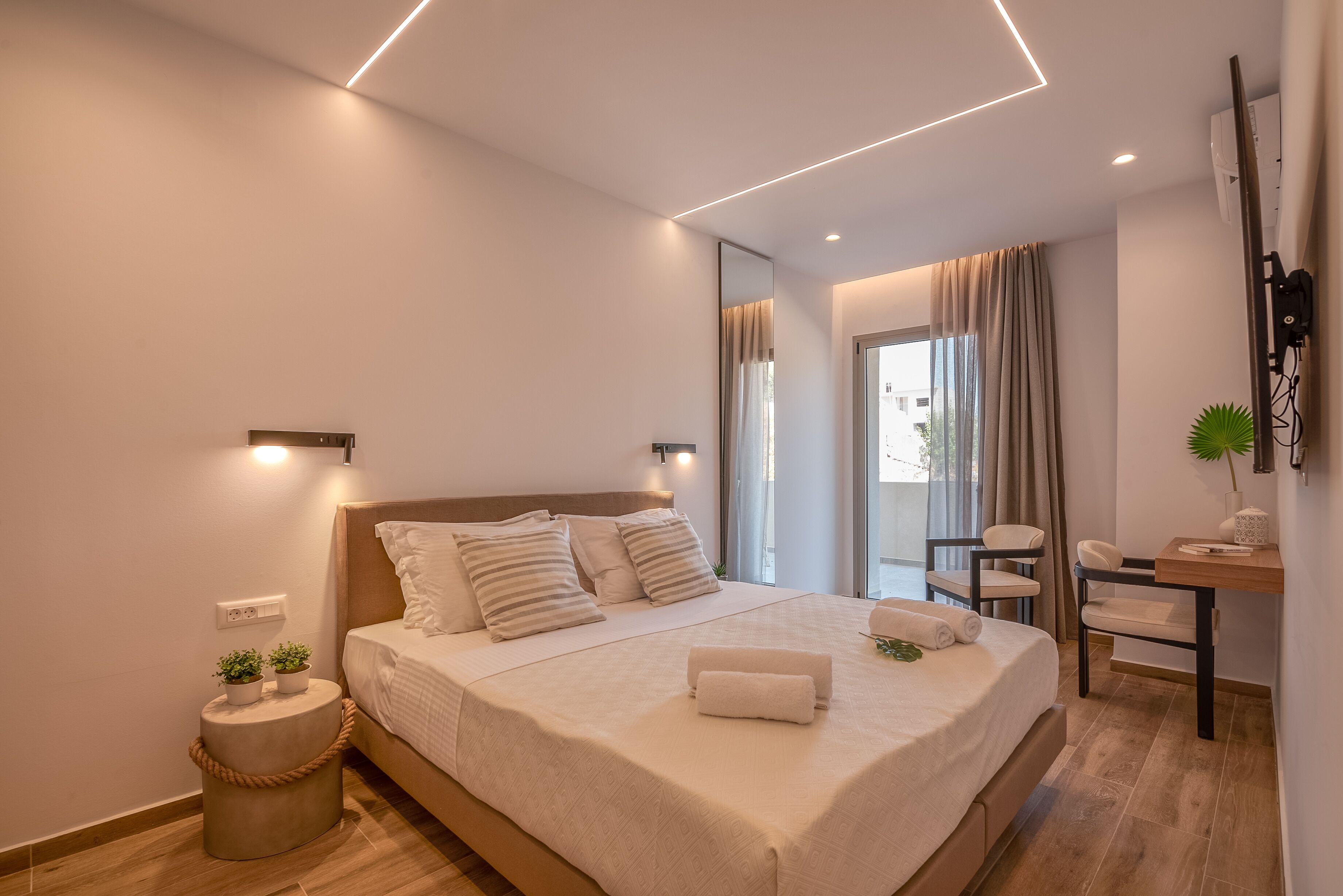 Quarto moderno com uma cama grande, mesas de cabeceira, uma cadeira e uma pequena mesa com uma planta.