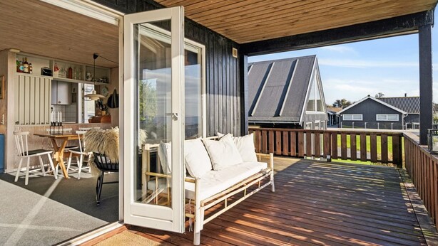 Una terraza de madera con sofá y sillas, un ventanal de vidrio que da a una cocina y vistas a casas vecinas.