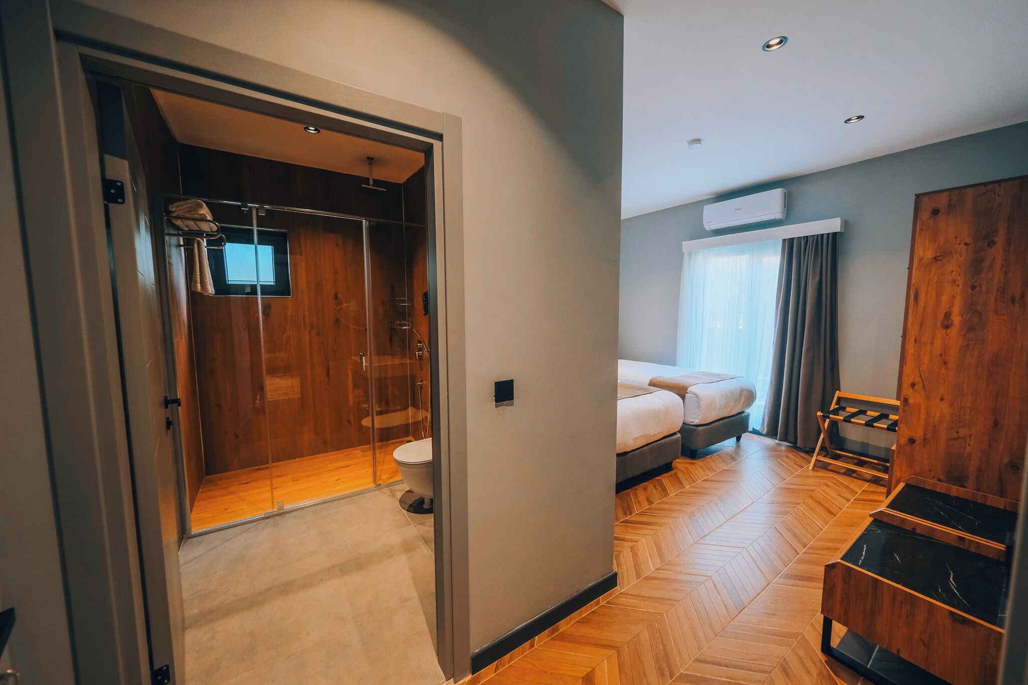 Quarto de hotel com piso de madeira, uma cama, guarda-roupa, banheiro com chuveiro e janela com cortinas.
