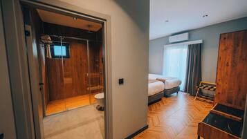Quarto de hotel com piso de madeira, uma cama, guarda-roupa, banheiro com chuveiro e janela com cortinas.