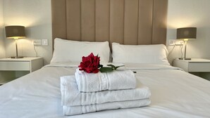 A neatly made bed with white linens, a red rose, and folded towels. There are two bedside lamps and a wall-mounted light fixture.