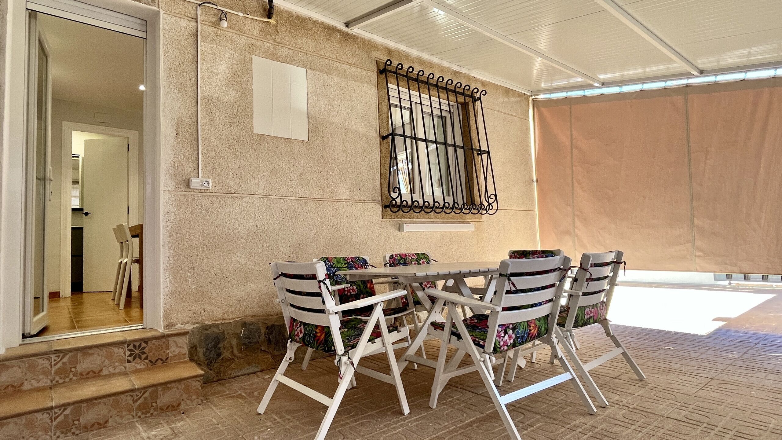 Un área de comedor al aire libre techada, con pared de piedra, una reja metálica y vista al interior.