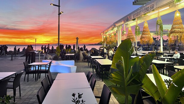 An outdoor restaurant with tables and chairs, overlooking a body of water during sunset.